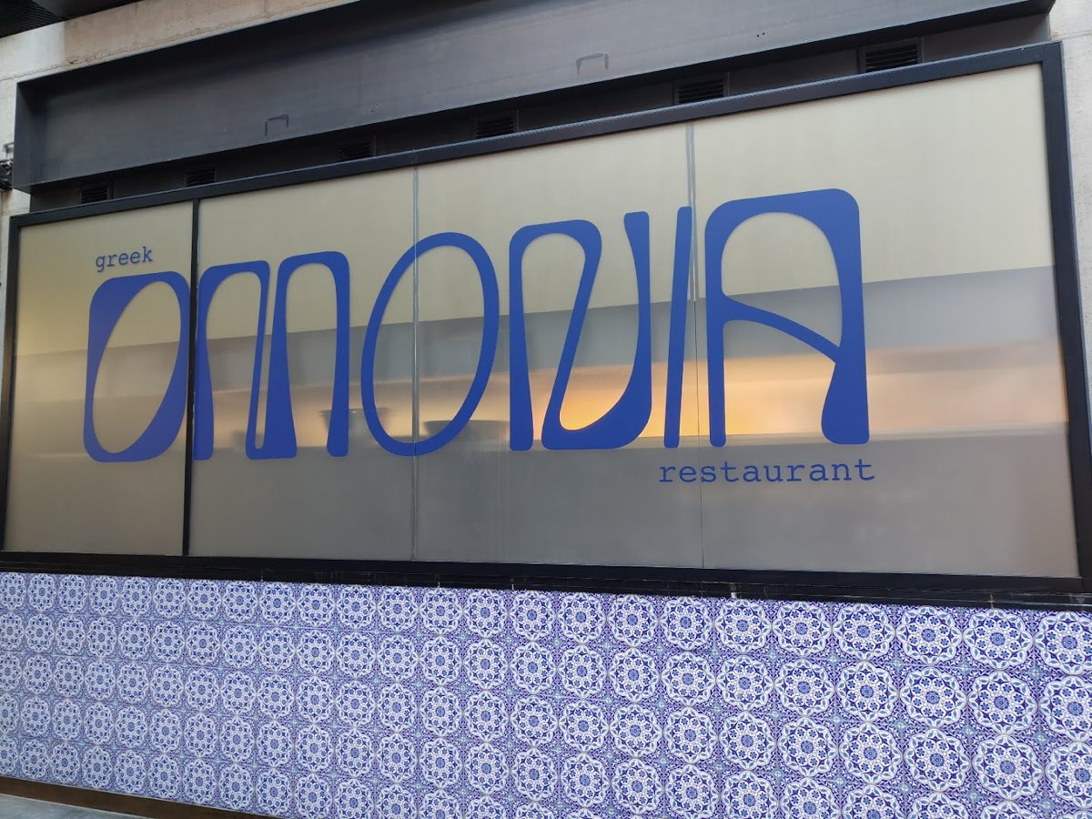 Kobaltowe logo Omonia na piaskowej ścianie nad niebiesko-białą grecką mozaiką